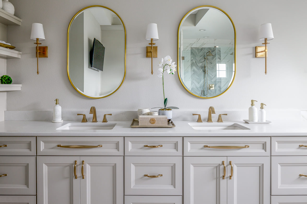 Double Sinks in Primary Bathroom with Gold Oval Mirrors and Wall Sconces Above Spacious Bathroom Cabinets | Compelling Homes Remodel + Design