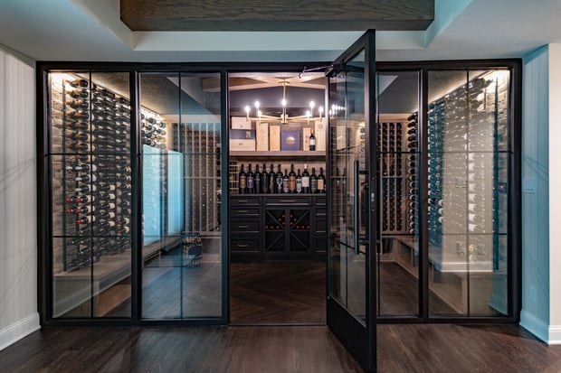 Glass-enclosed wine cellar with herringbone hardwood floor, black-framed glass doors, and dramatic chandelier