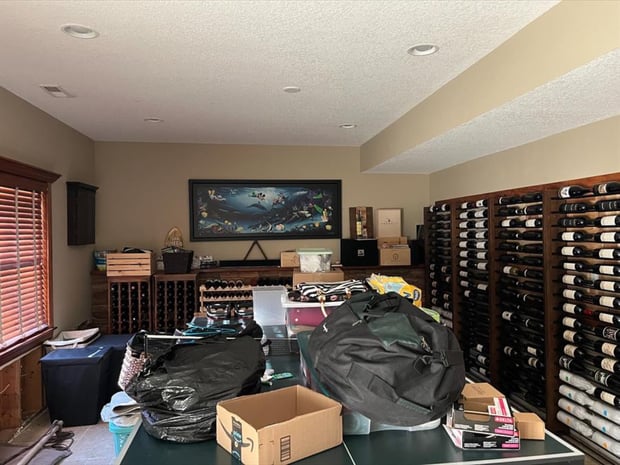 Cluttered basement storage area with bottle racks, mixed shelving, and recessed ceiling lights before remodel