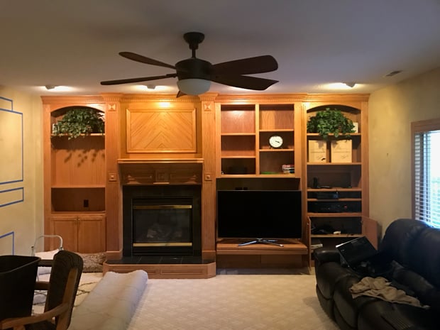 Builder-grade basement living room with oak built-in entertainment center, dated fireplace, and ceiling fan before remodel