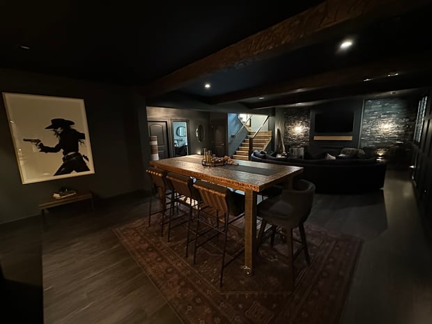 Dramatic dark basement with black walls, wood ceiling beams, stacked stone fireplace, and copper bar with stools