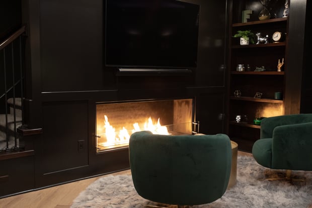 Dark moody basement with black walls, modern linear fireplace, mounted TV, and emerald velvet swivel chairs