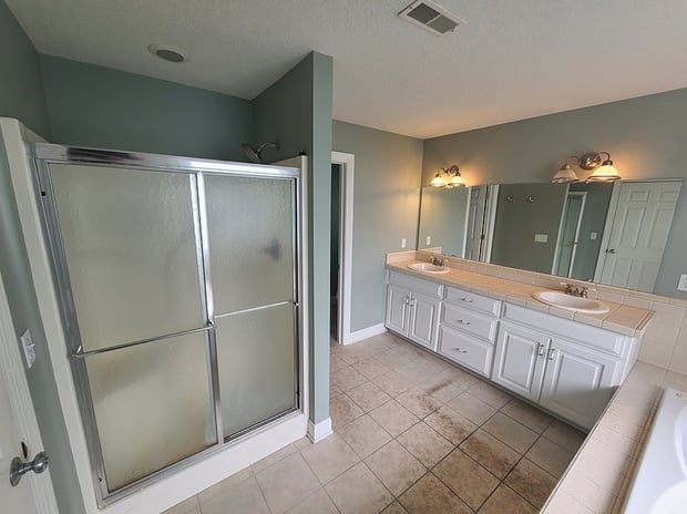 Outdated bathroom with sage walls, white double vanity, glass block shower, and dated tile floor before remodel