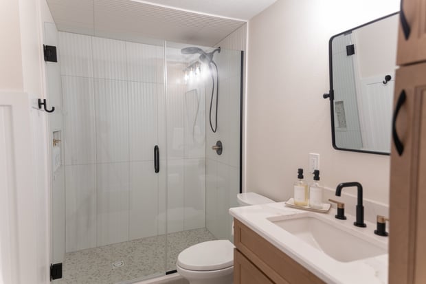 Modern hall bathroom with white shaker shower walls, oak vanity, brass fixtures, and pebble shower floor