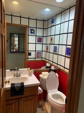 Quirky powder room with grid-pattern tile walls, red wainscoting, oak vanity, and eclectic art before remodel