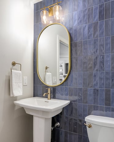 Modern powder room with blue subway tile feature wall, brass-framed mirror, brass sconces, and white pedestal sink