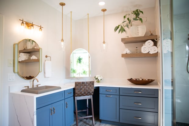 Modern bathroom with blue painted cabinets, brass pendant lights, marble counters, gold-framed mirror, and floating shelves