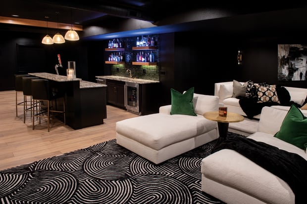 Modern dark basement bar with backlit liquor display, dark island, white sectional, and patterned rug