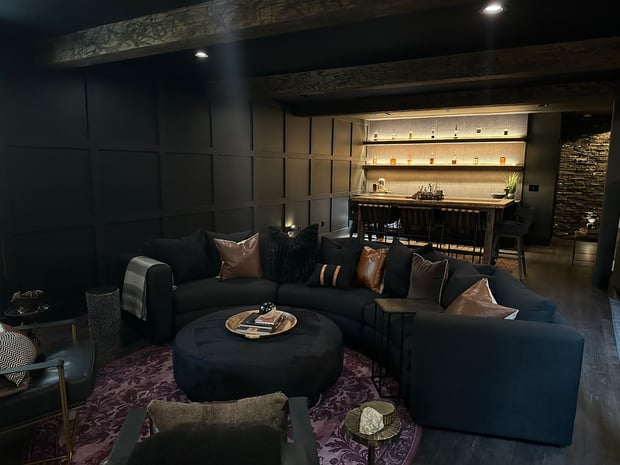 Dark moody basement living area with black panel walls, wood beam ceiling, and backlit liquor display
