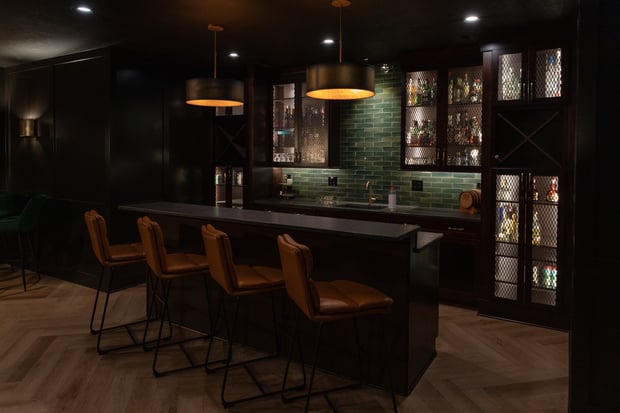 Modern basement bar with black cabinetry, green subway tile backsplash, leather stools, and herringbone wood floor