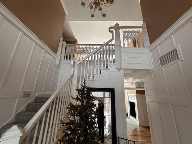 Traditional staircase with ornate scrollwork balusters, brown walls, and brass chandelier in two-story foyer before remodel