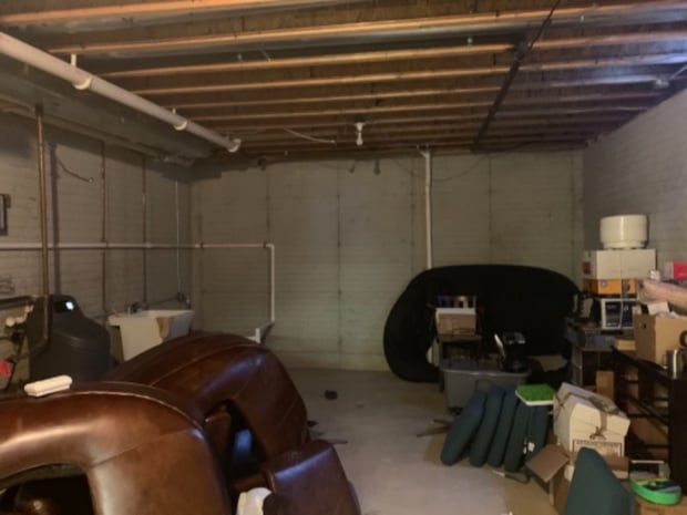 Unfinished basement with exposed ceiling joists, painted concrete walls, and stored furniture before remodel